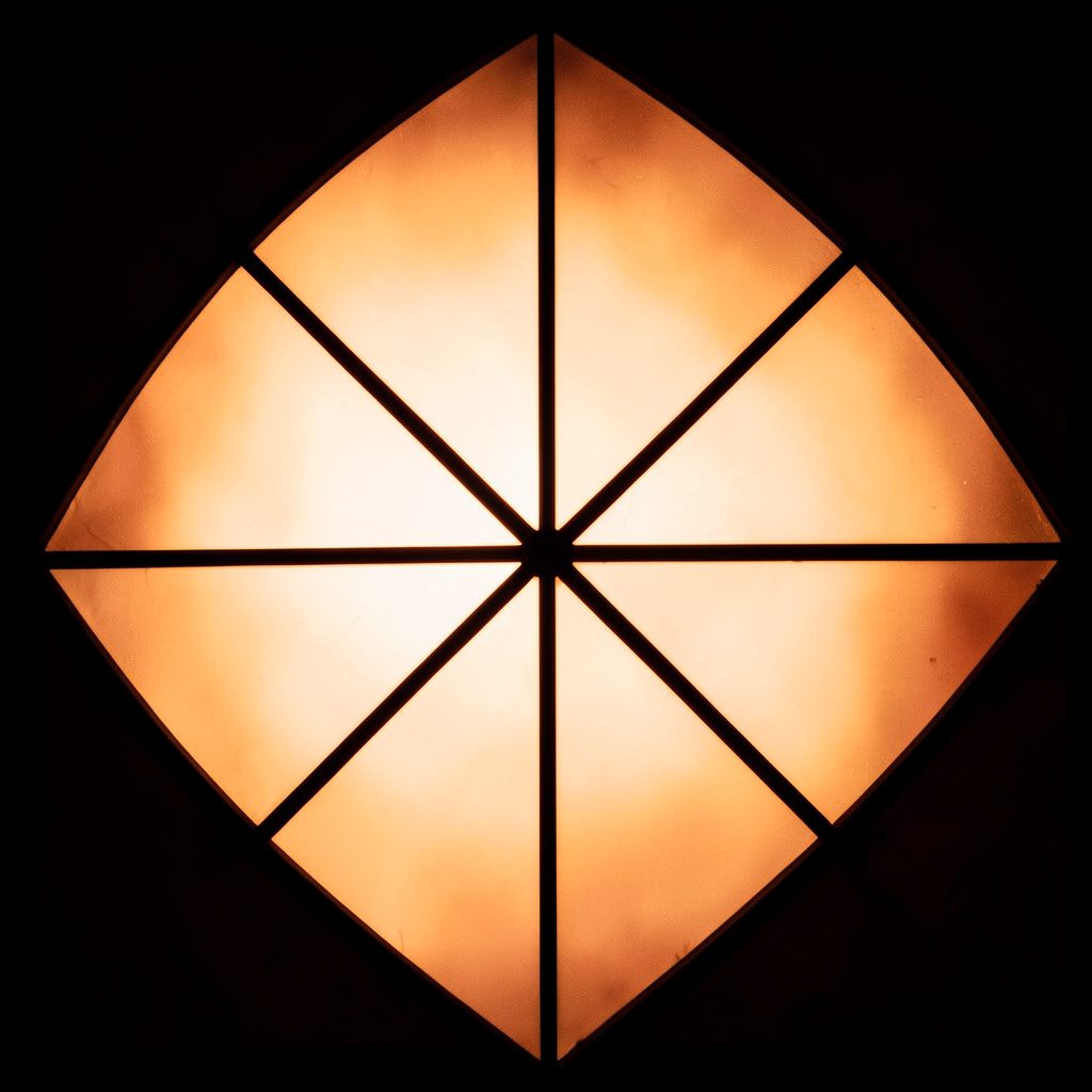 A stained-glass composition with bold leaded segments in deep amber, emerald green, and cobalt blue, forming an abstract geometric pattern that glows warmly against a dark background.